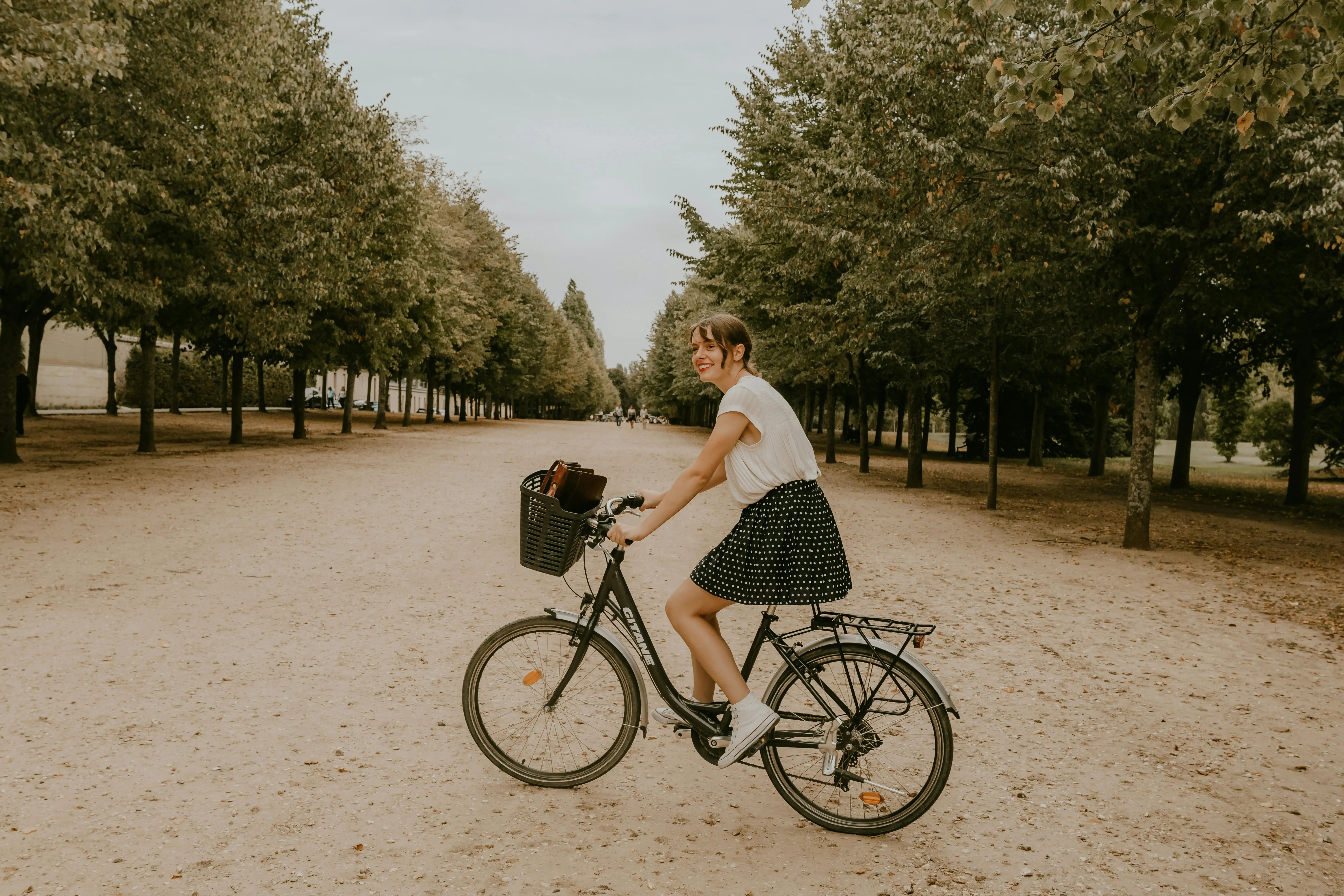 A joyful woman riding a bicycle along a tree-lined path in a Parisian park.